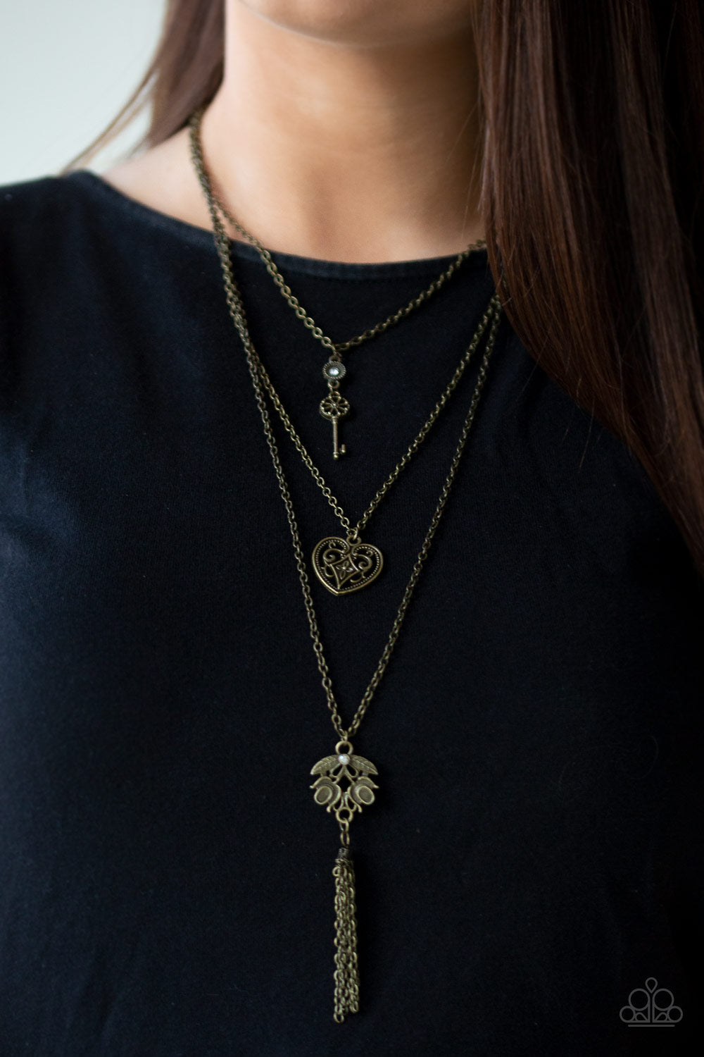 Paparazzi Necklace - A shimmery brass key, heart-like frame, and leafy brass pendant layer down the chest in a whimsical fashion. Infused with a playful tassel, glittery white rhinestones adorn the upper and lowermost frames for a sparkling finish.